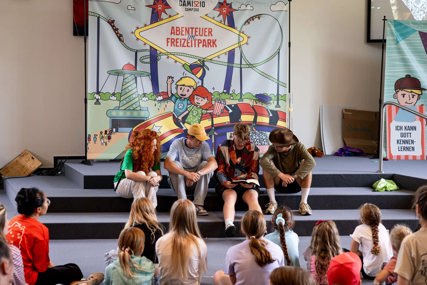 Vier Jugendliche sitzen vor einem bunten Banner mit der Aufschrift „Abenteuer im Freizeitpark“ und lesen gemeinsam in einem Buch, während Kinder zuschauen.
