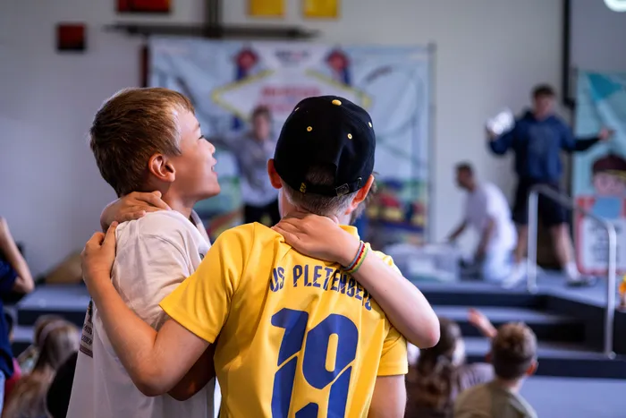 Zwei Jungen umarmen sich im Vordergrund vor einer Bühne mit weiteren Kindern.