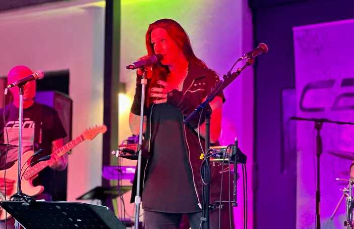 Sängerin der Band Cane singt bei ihrem Abschiedskonzert in ein Mikrofon, während im Hintergrund ein Gitarrist spielt.