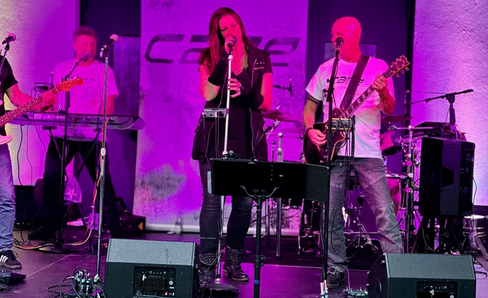 Die Band Cane spielt bei ihrem Abschiedskonzert auf einer Bühne unter lila Beleuchtung mit einer Sängerin, einem Gitarristen und einem Keyboarder.