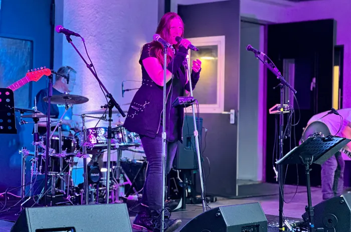 Eine Sängerin der Band Cane singt bei einem Abschiedskonzert unter violettem Bühnenlicht, im Hintergrund ist ein Schlagzeuger zu sehen.