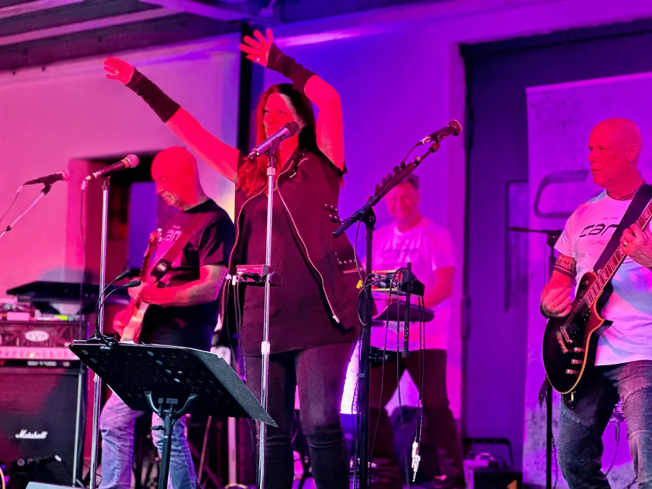 Sängerin mit erhobenen Armen und Bandmitglieder bei einem Live-Auftritt unter lila Bühnenlicht.