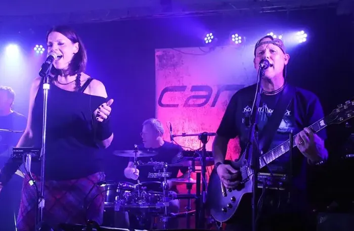 Die Band Cane tritt live auf einer Bühne unter violettem Licht auf; im Vordergrund singt eine Frau und ein Mann spielt Gitarre.