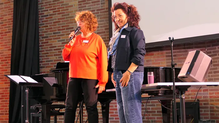 Ulrike Wahren spricht in ein Mikrofon, während eine Frau in einer dunklen Jacke neben ihr auf einer Bühne vor einem Klavier steht.