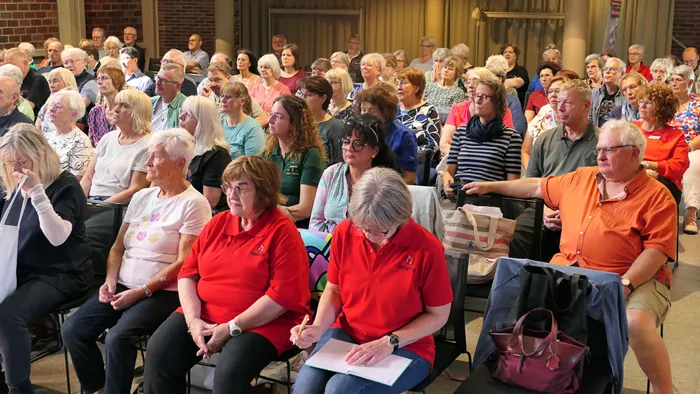 Eine große Gruppe von Menschen sitzt in Reihen in einem Saal während eines Chorcoachings.