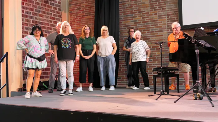 Teilnehmer eines Chorcoachings stehen auf einer Bühne vor einer Ziegelwand, rechts steht ein Mann an einem Flügel.