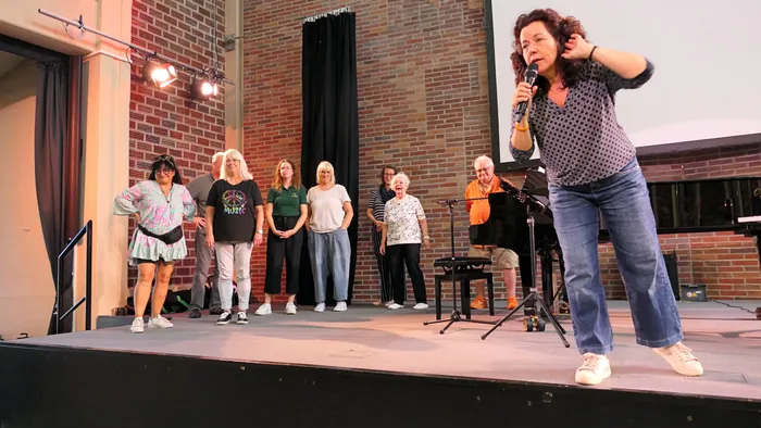 Ulrike Wahren spricht mit einem Mikrofon zu einer Gruppe von Chorsängern auf einer Bühne vor einer Ziegelwand.