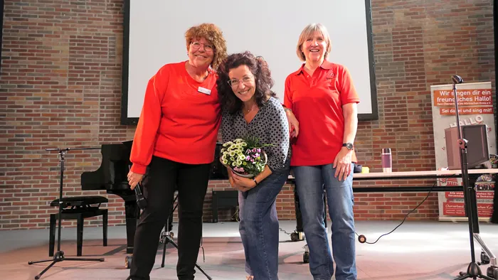Drei lächelnde Frauen stehen auf einer Bühne vor einer Leinwand und einem Flügel; die mittlere Frau hält einen Blumenstrauß.