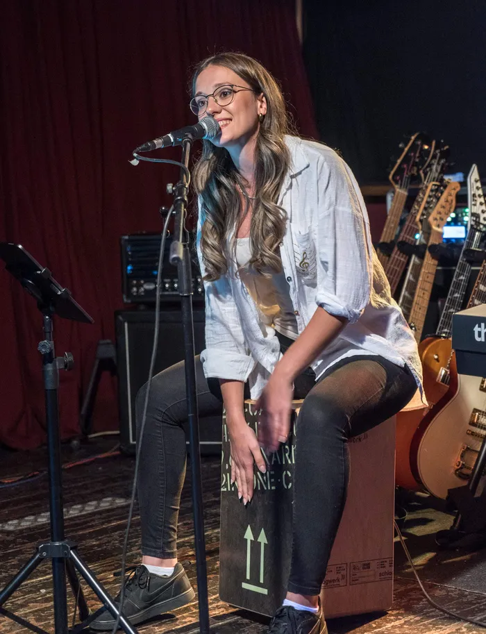 Eine junge Frau mit Brille singt in ein Mikrofon und sitzt auf einem Cajón beim Benefizkonzert „Rock for Peace“.