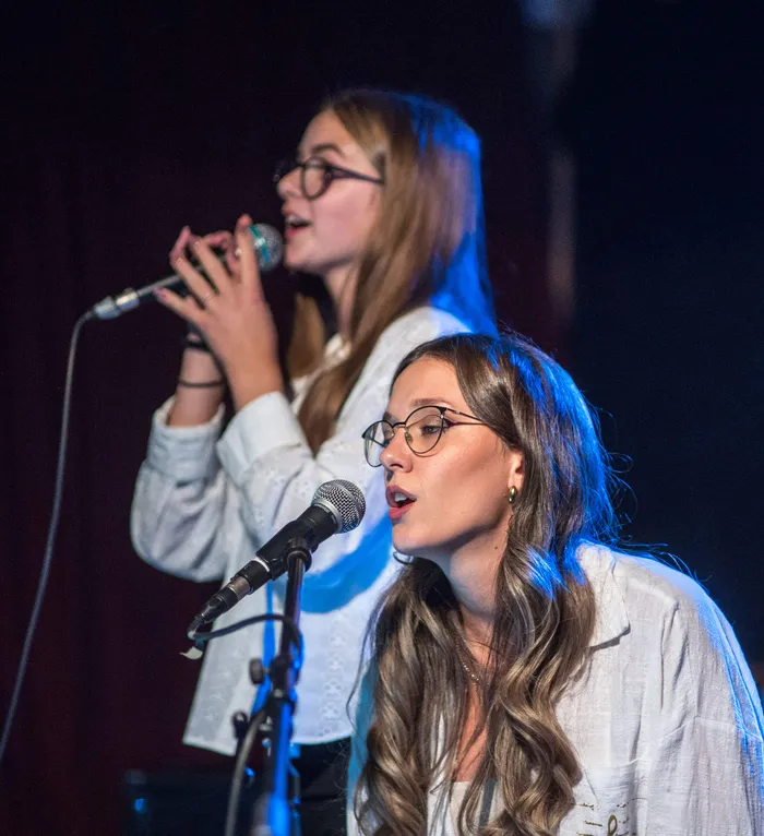Zwei junge Frauen mit Brillen und weißen Hemden singen bei einem Benefizkonzert in Mikrofone.