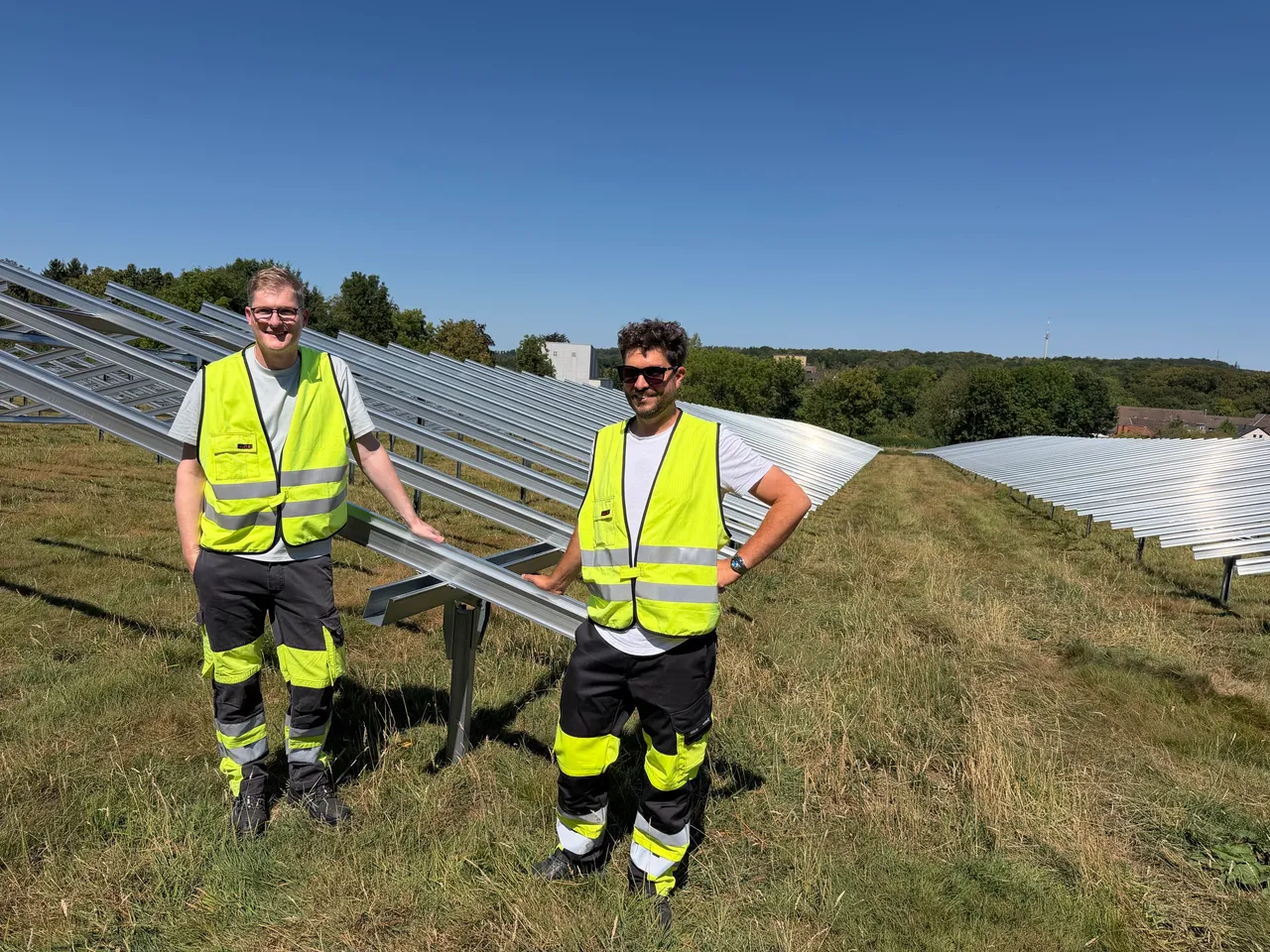 Zwei Männer in gelben Warnwesten und Arbeitskleidung stehen vor den Metallgestellen einer Freiflächen-Photovoltaikanlage auf einer Wiese.