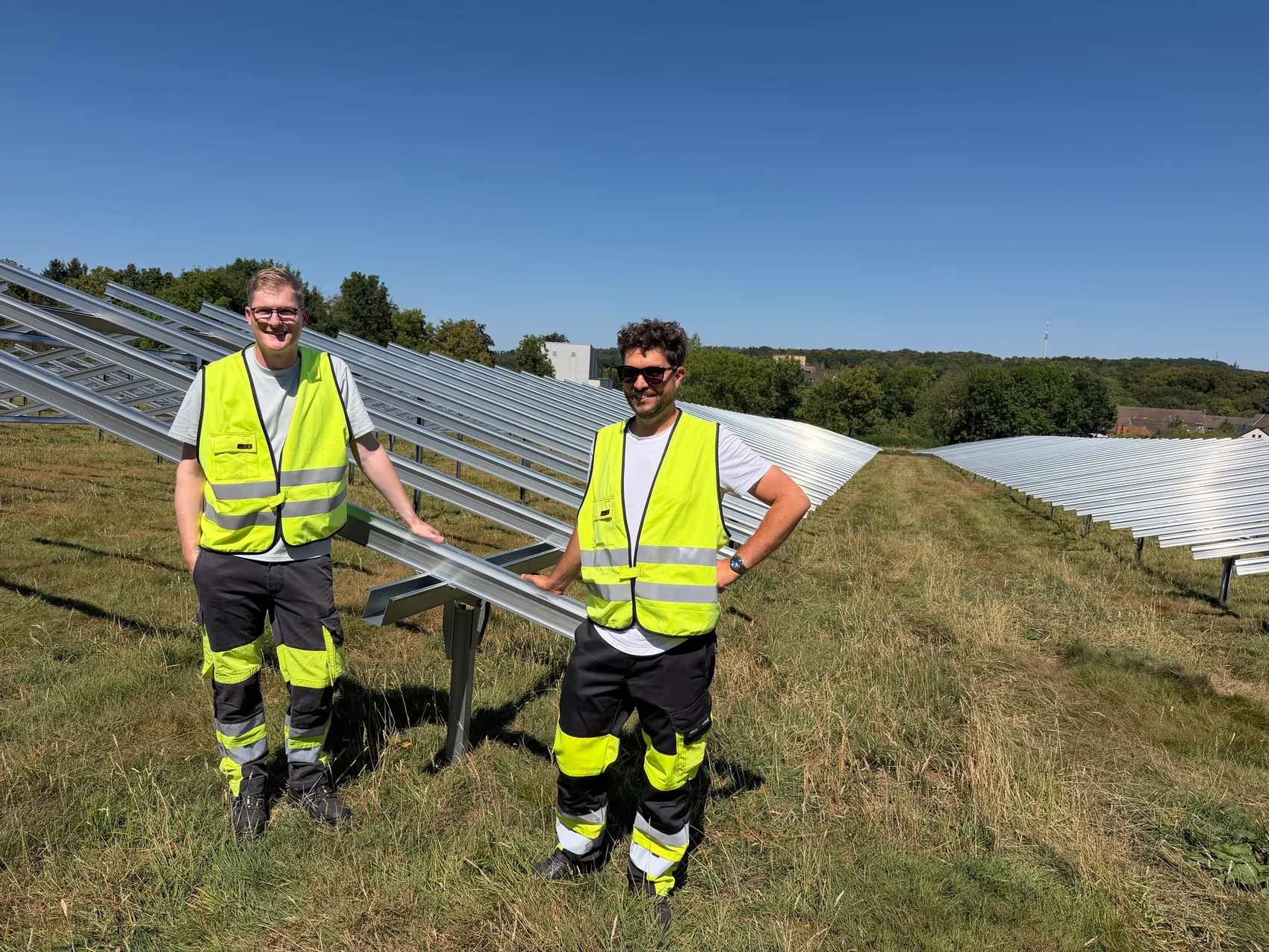 Zwei Männer in gelben Warnwesten und Arbeitskleidung stehen vor den Metallgestellen einer Freiflächen-Photovoltaikanlage auf einer Wiese.