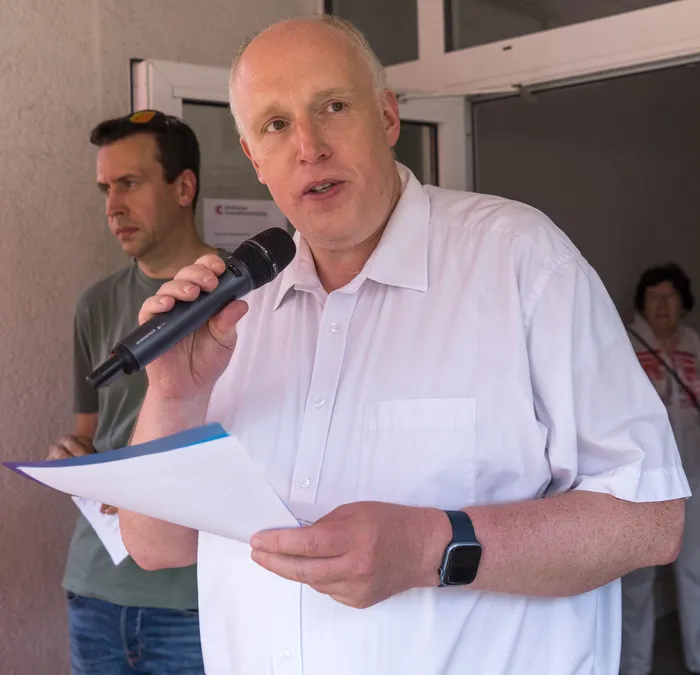 Ein Mann mit Glatze spricht in ein Handmikrofon, während er Notizen in der Hand hält. Im Hintergrund sind weitere Personen und ein Gebäude zu sehen.