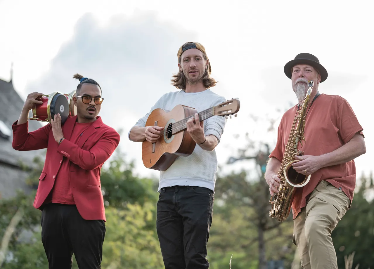 Drei Musiker spielen im Freien Trommel, Akustikgitarre und Saxophon.