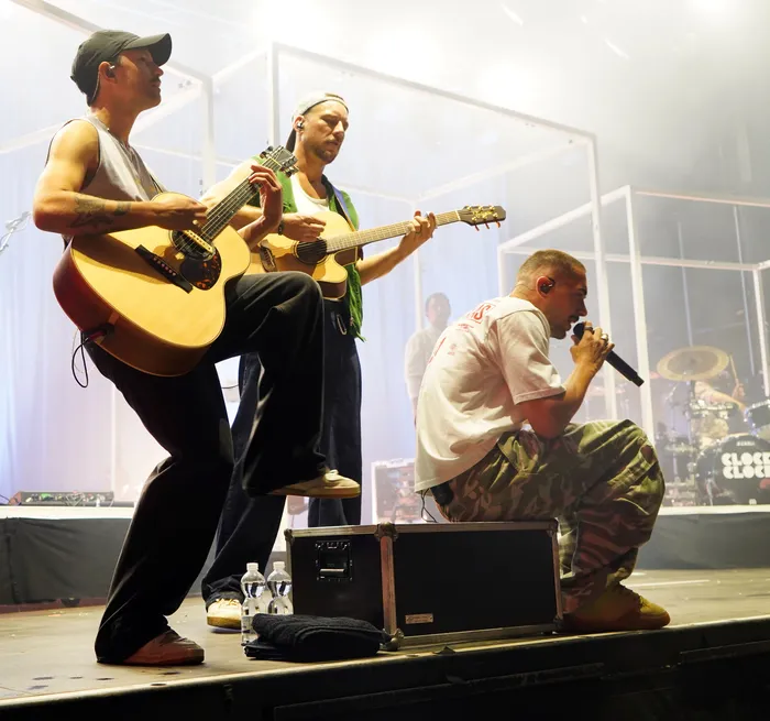 Zwei Musiker spielen Akustikgitarren, während ein Sänger im Vordergrund kniet und in ein Mikrofon singt.