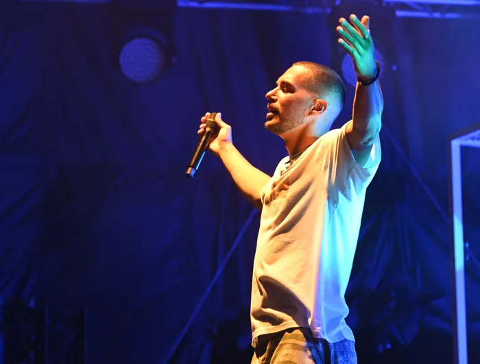 Ein Mann mit Bart und weißem T-Shirt singt mit einem Mikrofon auf einer blau beleuchteten Bühne.