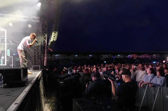 Ein Sänger in weißem T-Shirt und Camouflage-Hose singt auf einer beleuchteten Bühne vor einer großen Menschenmenge bei einem nächtlichen Konzert.