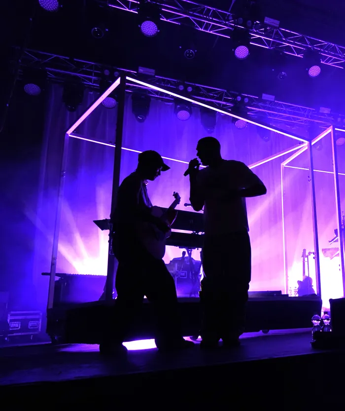 Silhouetten der Band Clockclock auf einer Bühne unter violettem Licht, während ein Musiker Gitarre spielt und der andere singt.
