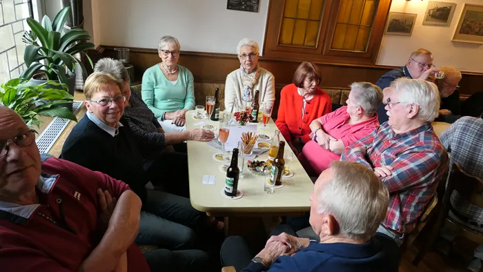 Eine Gruppe von Menschen sitzt um einen Tisch in einer Kneipe, Getränke vor sich. Im Vordergrund lächelt eine Frau in einem blau-weiß gestreiften Oberteil. Im Hintergrund ist ein Mann mit rotem Jackett zu sehen.