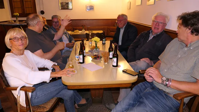 Eine Gruppe von sechs Personen sitzt um einen Tisch in einer Kneipe, Getränke und Snacks davor. Eine Frau hält ein Glas hoch, ein Mann hebt die Hand. Im Hintergrund sind weitere Personen und Dekorationen zu sehen.