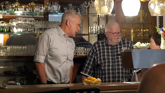 Zwei Männer stehen an einer Bar, der eine mit hochgekrempelten Ärmeln und der andere mit kariertem Hemd. Im Hintergrund eine Vitrine mit Pokalen und Gläsern.