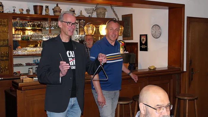 Ein Mann mit Brille und im Anzug hält einen Tamburin, während ein anderer Mann in einem gestreiften Trikot einen Dreieckschal schwingt. Im Hintergrund steht ein Mann mit Bart und eine Bar mit zahlreichen Flaschen und Dekorationen.