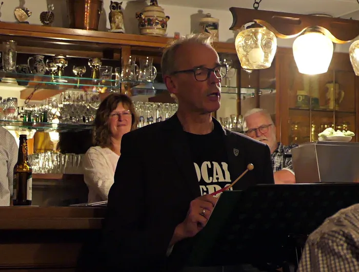 Ein Mann mit Brille und schwarzem Jackett spricht in eine Gruppe von Menschen in einer Kneipe. Im Hintergrund sind Regale mit Gläsern und Flaschen zu sehen.