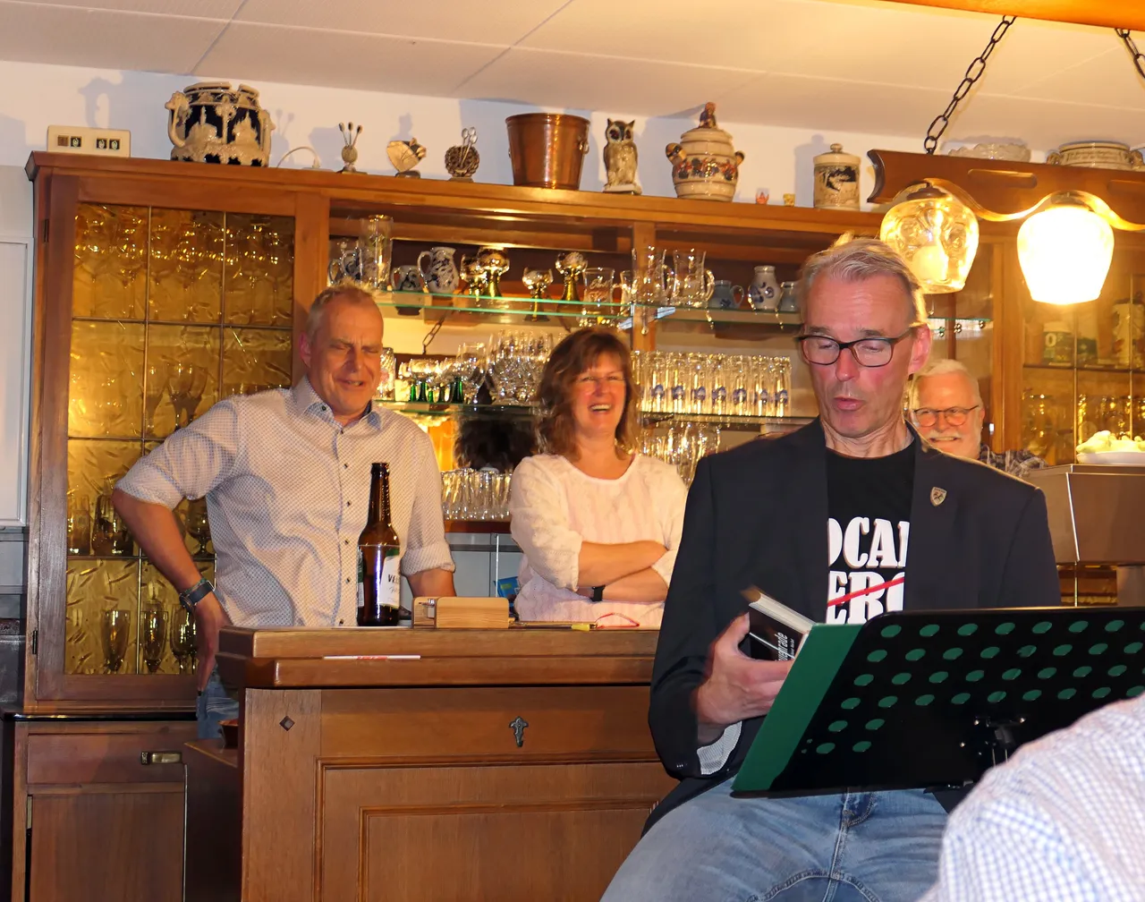 Ein Mann mit Brille liest von Notenblättern vor, während er in einer Kneipe steht. Im Hintergrund stehen und sitzen weitere Personen an einer Theke, über der eine Sammlung von Keramik und Gläsern ausgestellt ist.