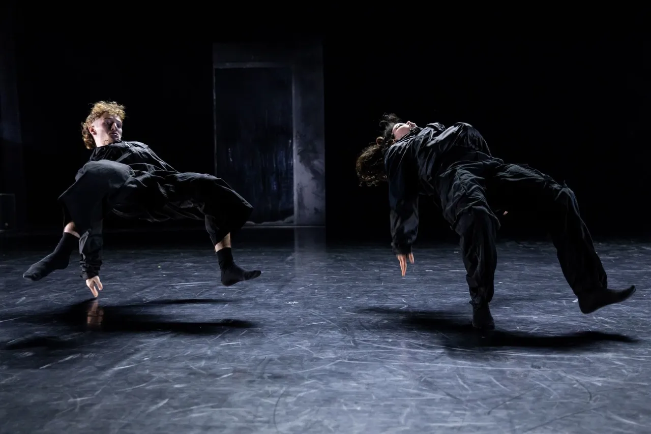 Zwei Tänzer in schwarzer Kleidung bei einer dynamischen Tanzperformance auf einer dunklen Bühne.