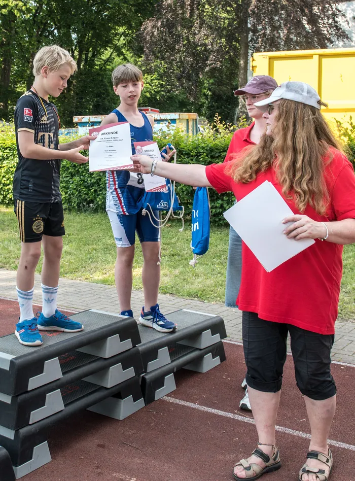 A young athlete in a blue and white sports outfit receives a certificate from a woman with a cap and red shirt. Another boy in a football jersey stands to the left, watching the exchange.