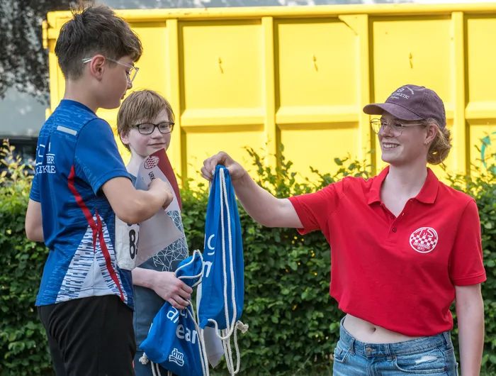 Eine Frau in rotem Poloshirt und Kappe überreicht einem Jungen mit Rucksack und einem weiteren Jungen mit Brille eine blaue Tasche mit Aufschrift. Im Hintergrund eine gelbe Containerwand.