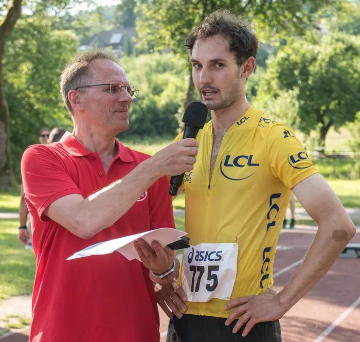 Ein Mann im gelben Trikot mit der Startnummer 175 wird von einem Reporter mit Mikrofon interviewt. Im Hintergrund sind Bäume und eine Laufstrecke zu sehen.