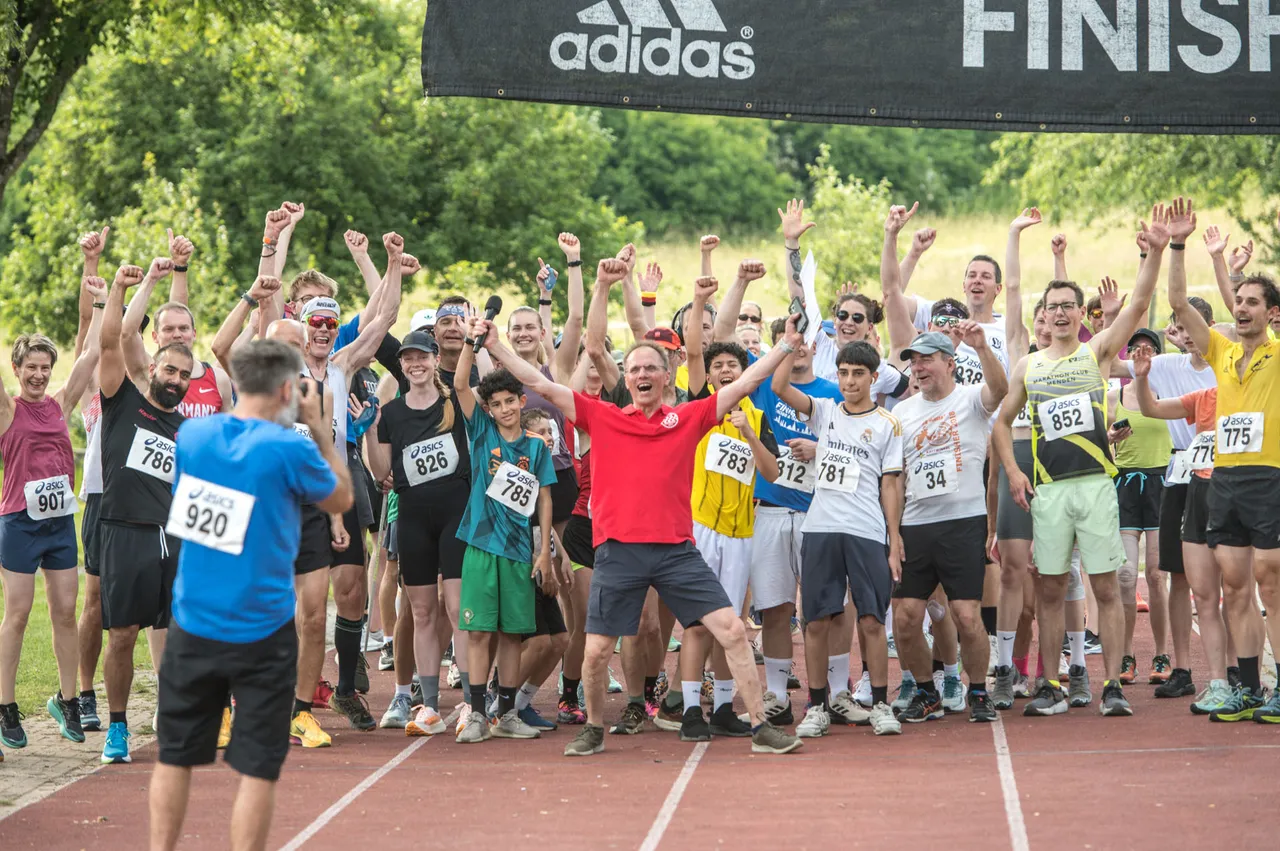 Eine Gruppe von Läufern steht mit erhobenen Armen auf einer roten Laufbahn vor einer Ziellinie mit dem Adidas-Logo. Viele tragen Startnummern und Sportkleidung.