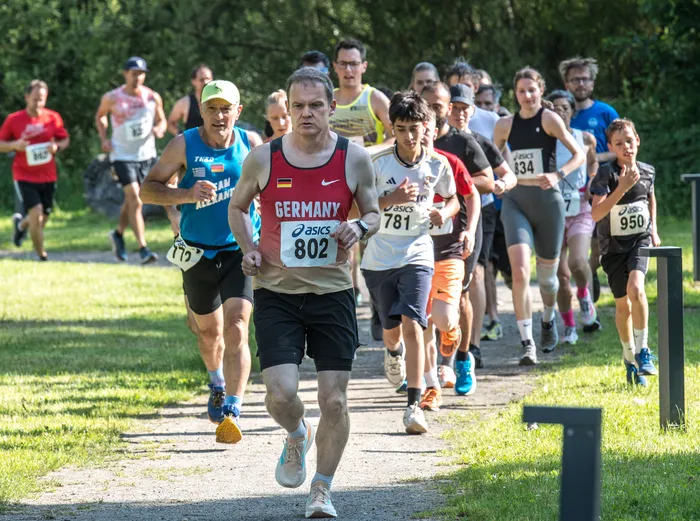 Ein Läufer mit der Nummer 802 und dem deutschen Wappen auf dem Trikot führt eine Gruppe von Teilnehmern bei einem Crosslauf an. Im Hintergrund sind weitere Läufer und Bäume zu sehen.