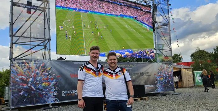 Zwei Männer im Trikot der deutschen Nationalmannschaft stehen vor einer großen Leinwand, auf der ein Fußballspiel gezeigt wird. Im Hintergrund ist ein Festivalgelände mit weiteren Zuschauern und Zelten zu sehen.