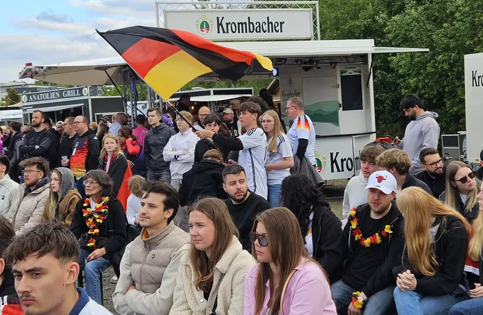 Eine Menschenmenge jubelt vor einer Bühne, viele tragen deutsche Flaggen oder schwarz-rot-goldene Kleidung und Girlanden. Im Hintergrund ist ein Krombacher-Stand zu sehen.
