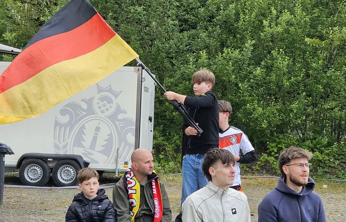 Ein Junge steht auf einem Anhänger und schwenkt eine große Deutschlandflagge. Im Vordergrund stehen jubelnde Fans, einige mit Deutschland-Schals.