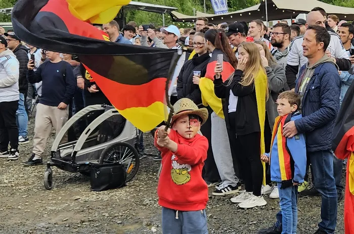 Junge mit rot-schwarzen Gesichtsbemalungen und Strohhut steht im Vordergrund und streckt die Arme aus, während im Hintergrund eine jubelnde Menschenmenge mit Deutschlandflaggen zu sehen ist.