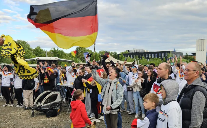 Jubelnde Menschenmenge mit Deutschlandflaggen feiert den Sieg der deutschen Nationalmannschaft gegen Curaçao bei einer Public-Viewing-Veranstaltung. Im Hintergrund ein Gebäude und ein Parkplatz.