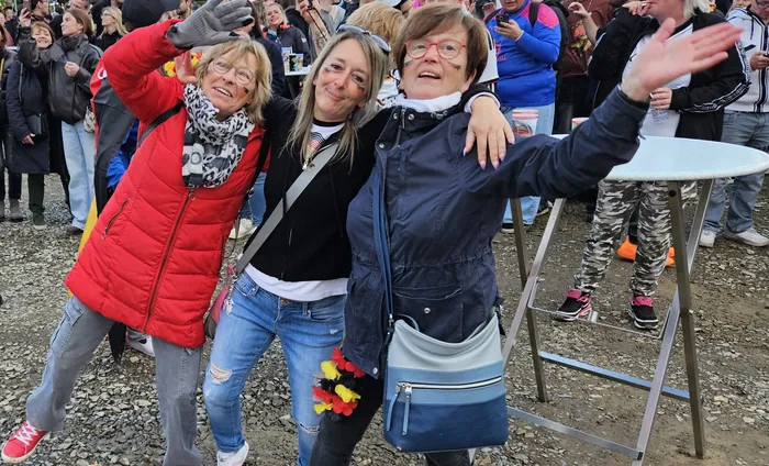 Drei Frauen umarmen sich und lächeln in die Kamera, im Hintergrund eine jubelnde Menschenmenge. Eine der Frauen trägt eine Deutschlandfahne am Handgelenk.