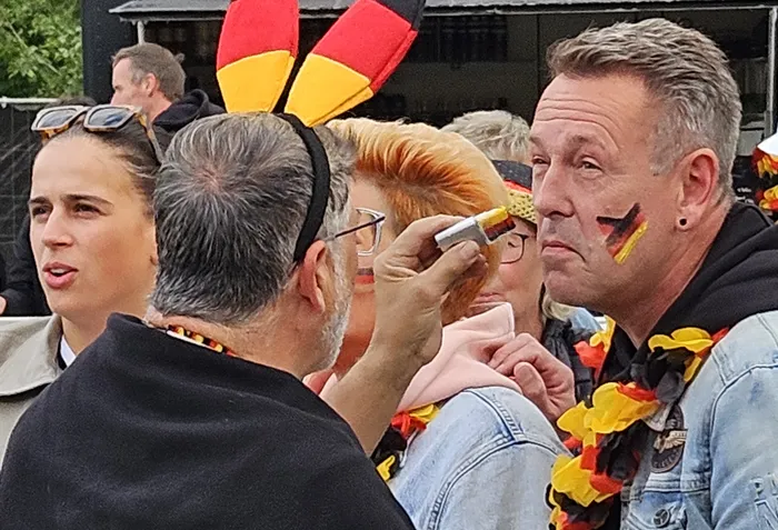 Ein Fan trägt eine Kopfbedeckung mit den deutschen Farben Schwarz, Rot und Gold und wird von einer Person mit Gesichtsbemalung in den gleichen Farben geschminkt. Im Hintergrund sind weitere Fans zu sehen, die die deutsche Nationalmannschaft unterstützen.