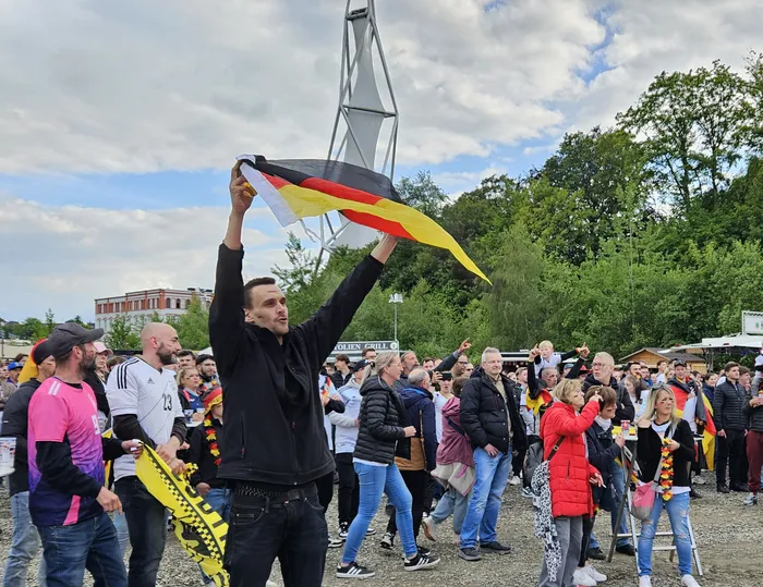 Ein Mann hält eine Deutschlandflagge in die Höhe, umgeben von jubelnden Fans auf einem Festivalgelände. Im Hintergrund sind weitere Menschen und Bäume zu sehen.