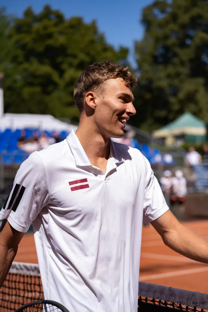 Kristians Koldvells lächelt auf einem Tennis-Sandplatz; er trägt ein weißes Poloshirt mit der lettischen Flagge.