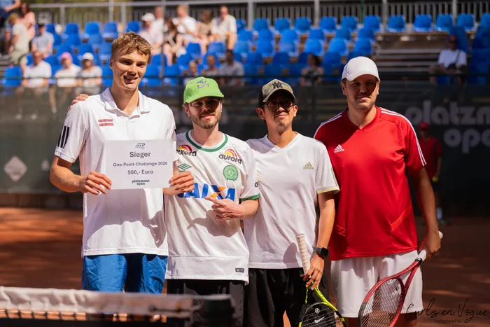 Vier Männer auf einem Tennisplatz, darunter Kristians Koldvells, der ein Schild mit der Aufschrift „Sieger One-Point-Challenge 2026 500,- Euro“ hält.
