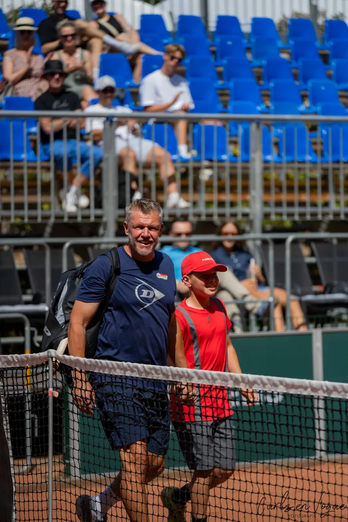 Ein Mann und ein Junge gehen auf einem Tennisplatz vor einem Netz, im Hintergrund sitzen Zuschauer auf blauen Tribünen.