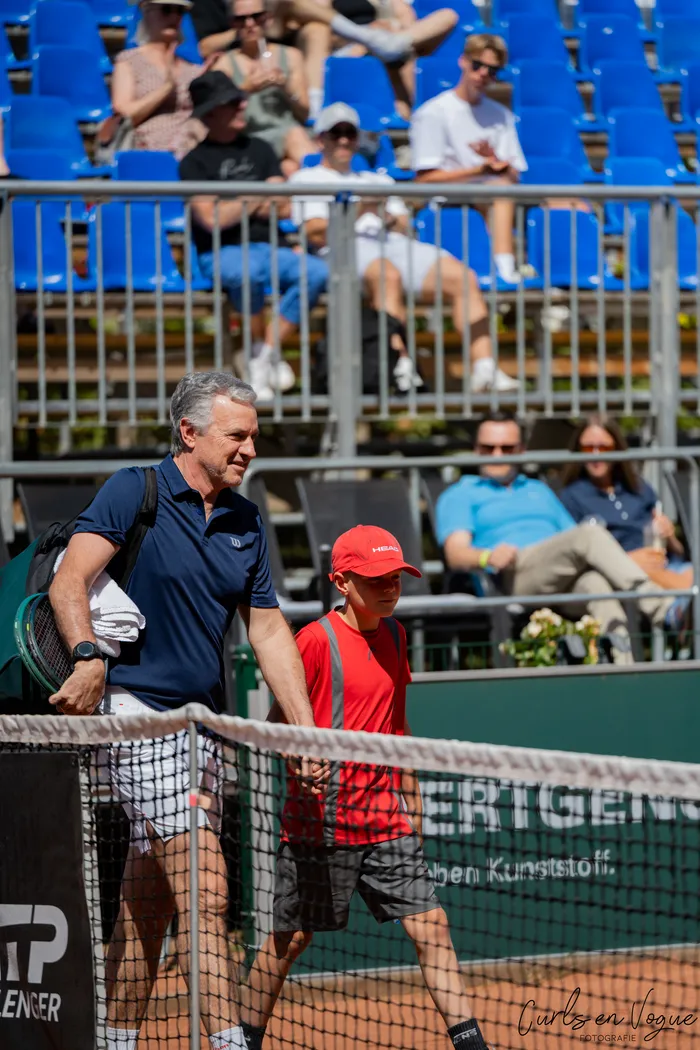 Ein Mann mit Tennistasche und Schläger geht neben einer Person in rotem T-Shirt und roter Kappe über einen Tennisplatz vor blauen Zuschauerrängen.