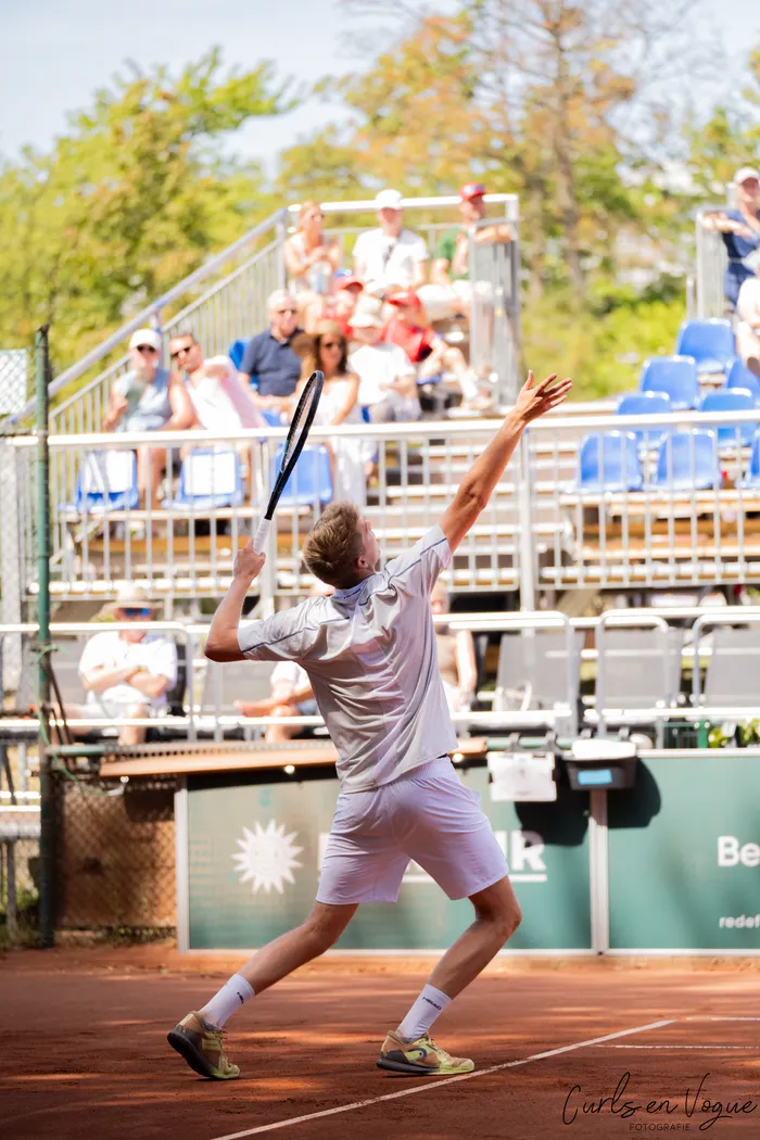 Ein Tennisspieler in weißer Sportkleidung führt einen Aufschlag auf einem Sandplatz vor Zuschauern aus.