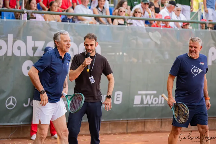Drei Männer bei einem Show-Match auf einem Tennisplatz; einer hält ein Mikrofon, die anderen beiden halten Tennisschläger und lächeln.