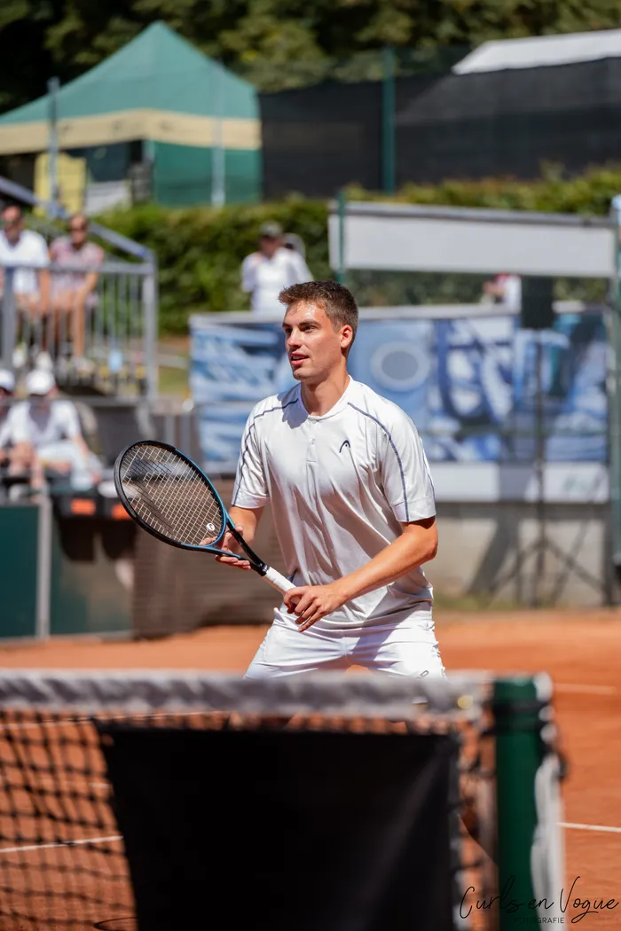 Ein Tennisspieler in weißer Sportkleidung hält seinen Schläger auf einem Sandplatz bereit.