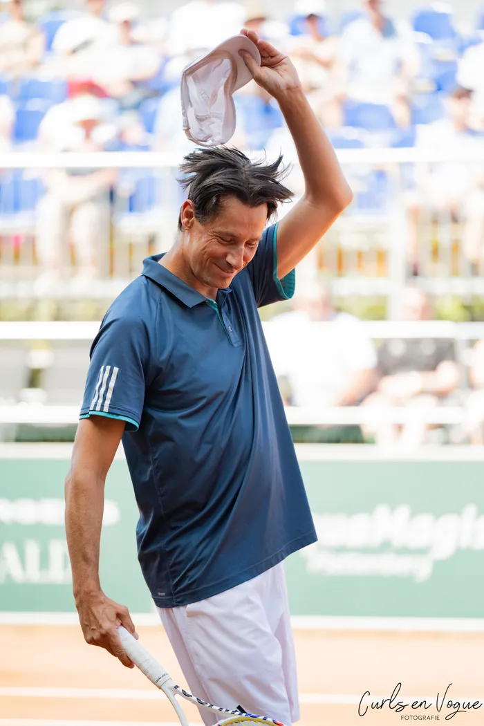 Ein lächelnder Tennisspieler in blauem Poloshirt hält eine weiße Kappe hoch und einen Tennisschläger in der Hand.